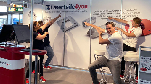 Vier Mitarbeiter und Mitarbeiterinnen am Laserteile4you-Stand zeigen auf die Messewand mit Logo